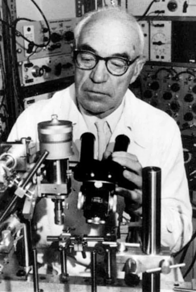 Foto em preto e branco de Sir John Carew Eccles, neurocientista ganhador do Prêmio Nobel, examinando um equipamento de laboratório.
