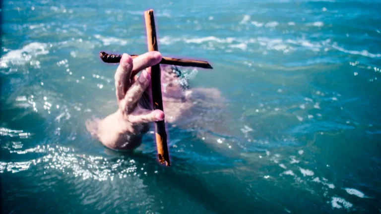 Una mano que emerge de las aguas turbulentas, sosteniendo una cruz de madera, simboliza un grito de salvación en los momentos finales de la vida.